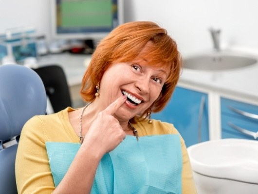 Older redheaded woman in dental chair pointing to her smile