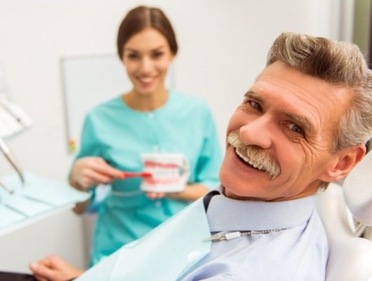 Senior man smiling in dental chair while dental assistant brushes dentures