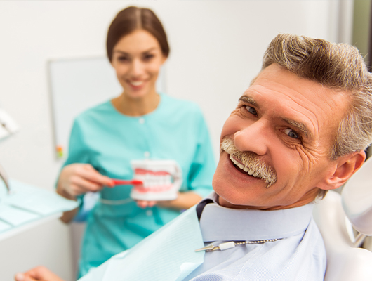 Smiling senior man in dental chair with dental assistant brushing dentures