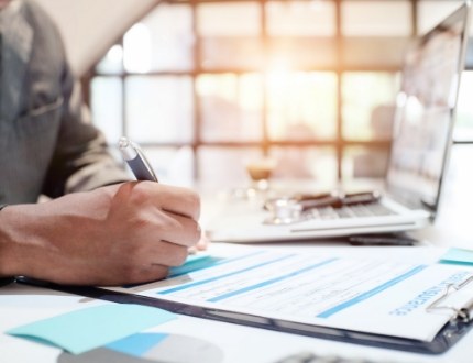 Person writing on form on clipboard while sitting at desk with laptop